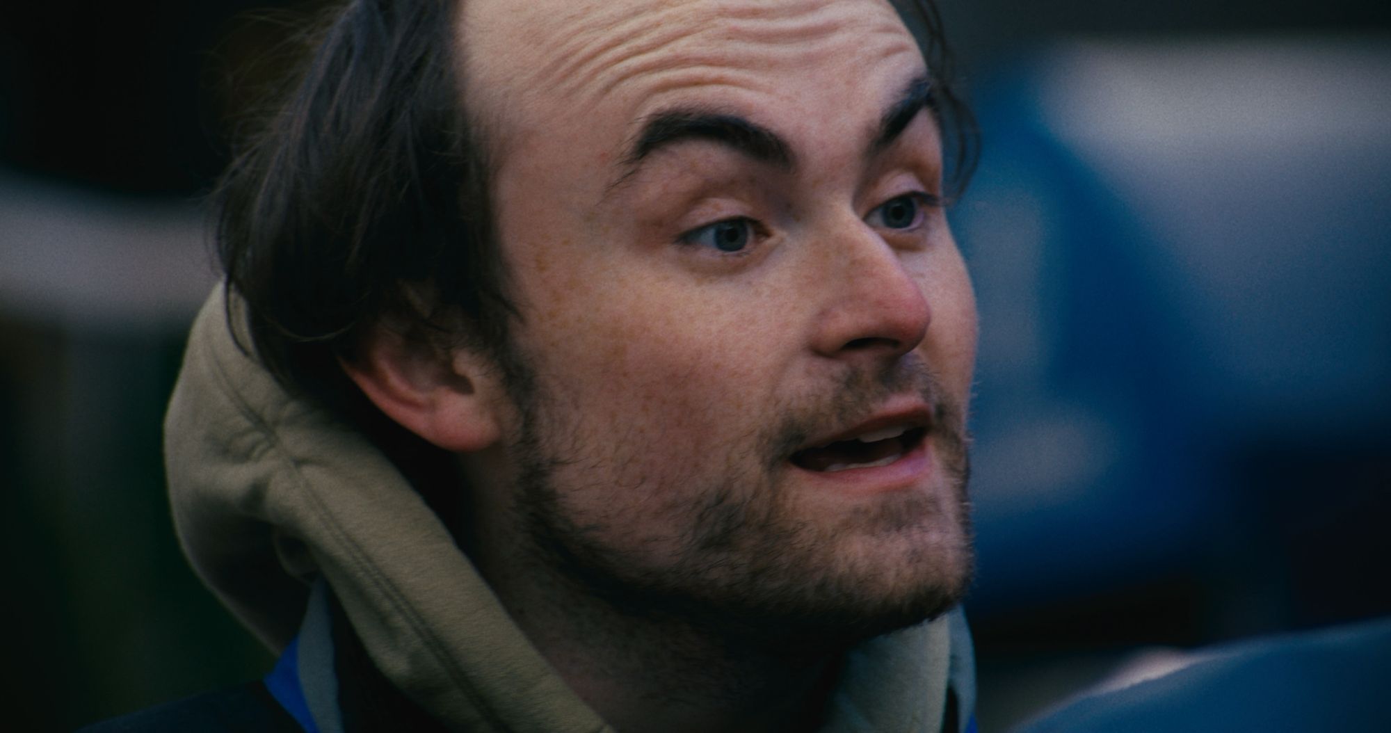 Close-up of a young man in a hooded jacket, mid-sentence.