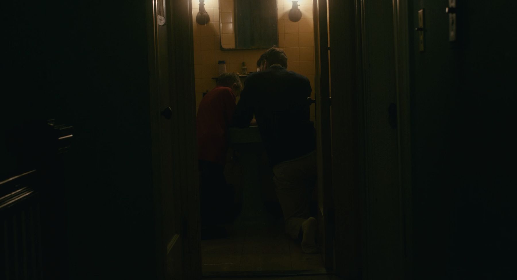 A father kneels beside his son at a bathroom sink, seen from a dark hallway.