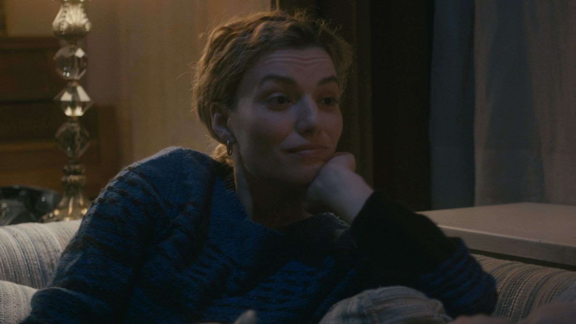 The mother smiles on the couch, chin on her hand.