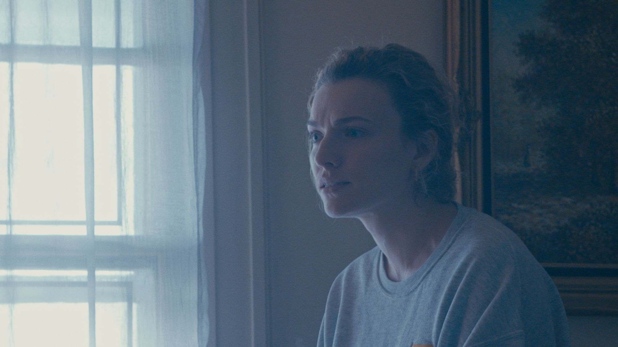 A mother in a grey sweatshirt sits by a curtained window in pale blue light.