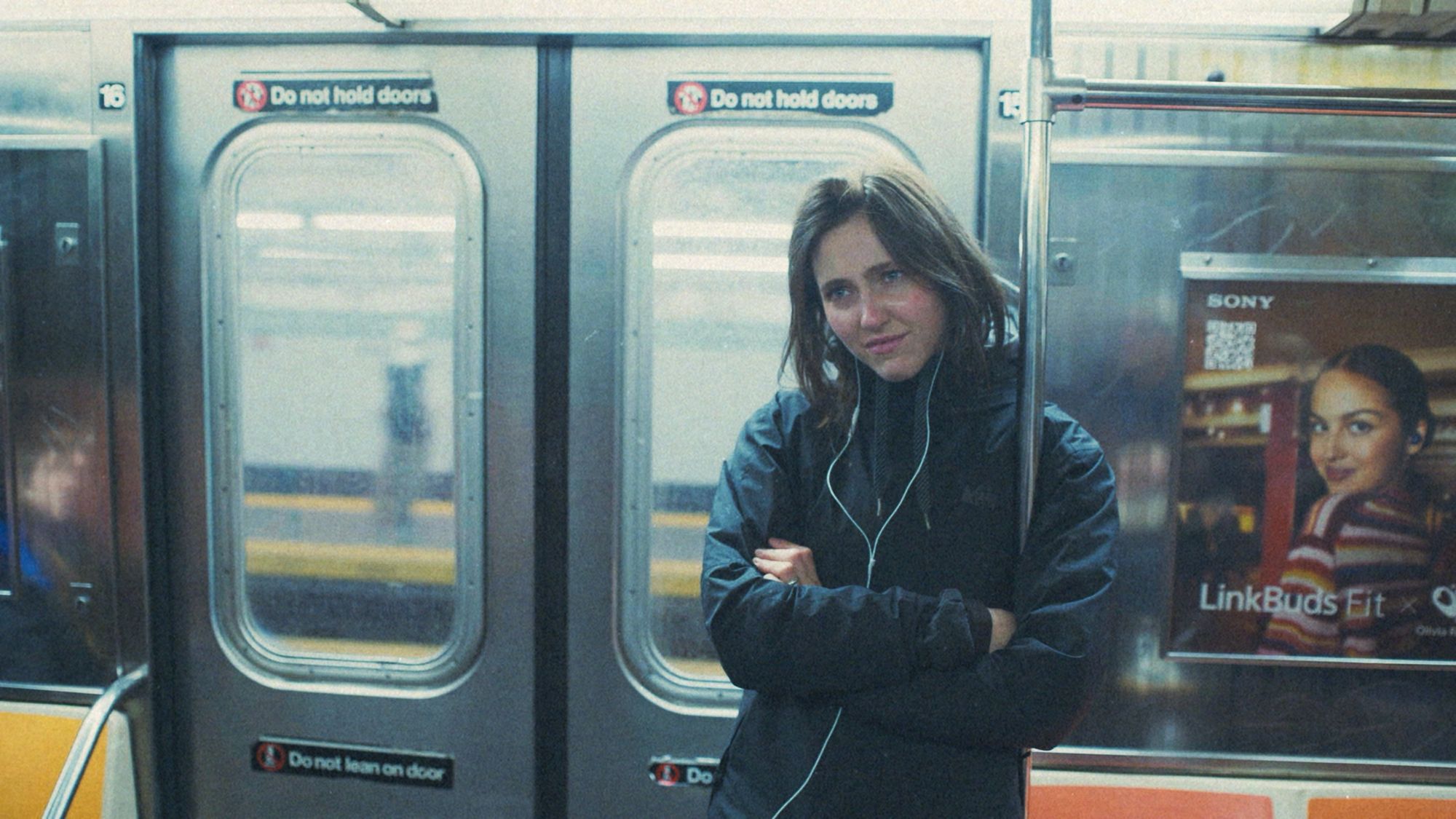 A young woman with earphones leans against subway doors, arms crossed.
