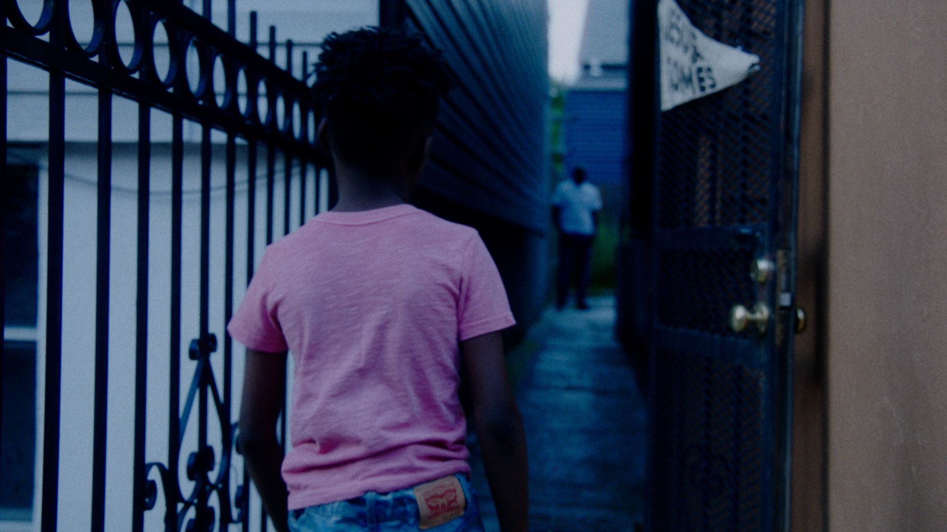 A boy in a pink shirt walks down a narrow blue alley toward a man.