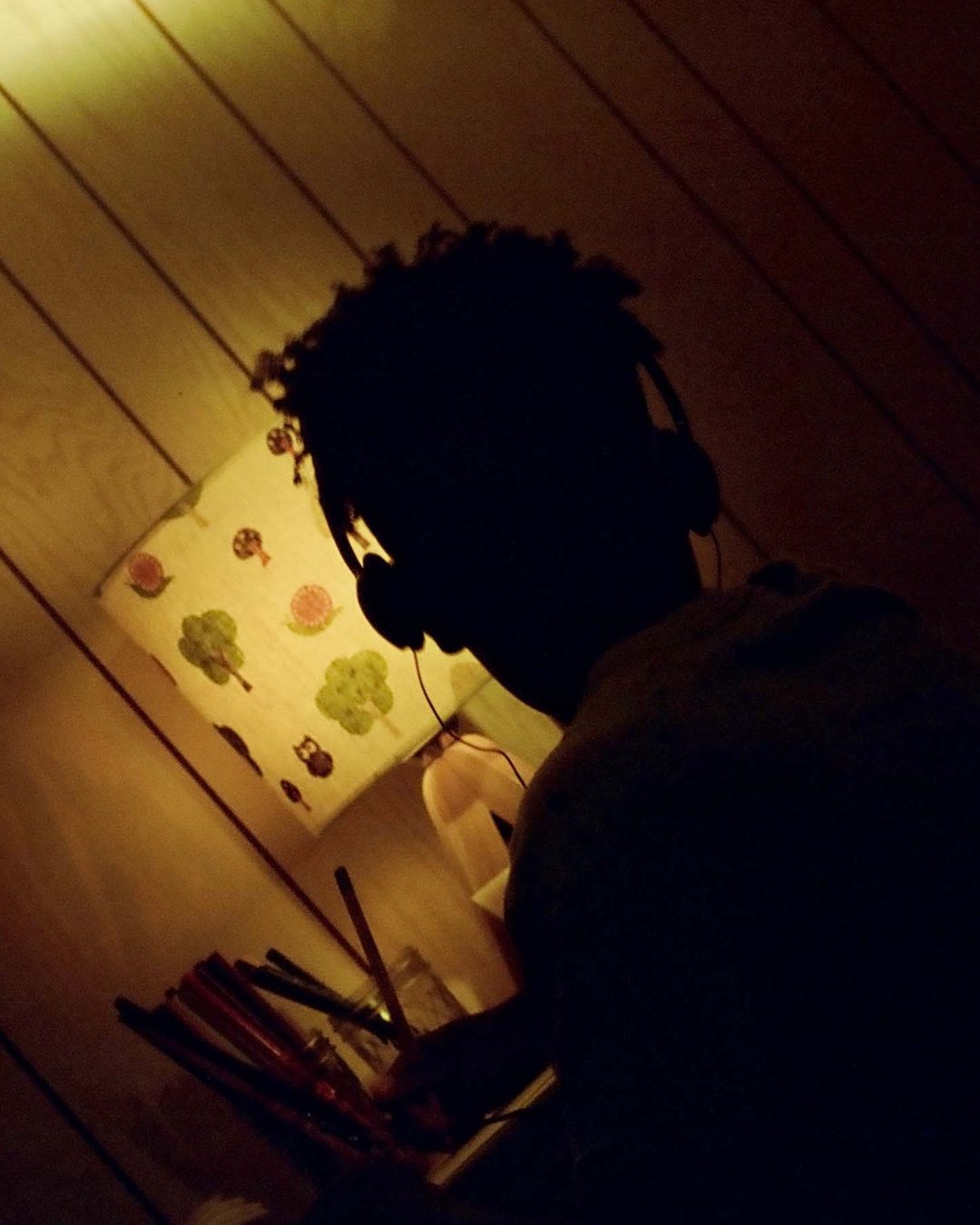 The boy draws by lamplight, headphones on.