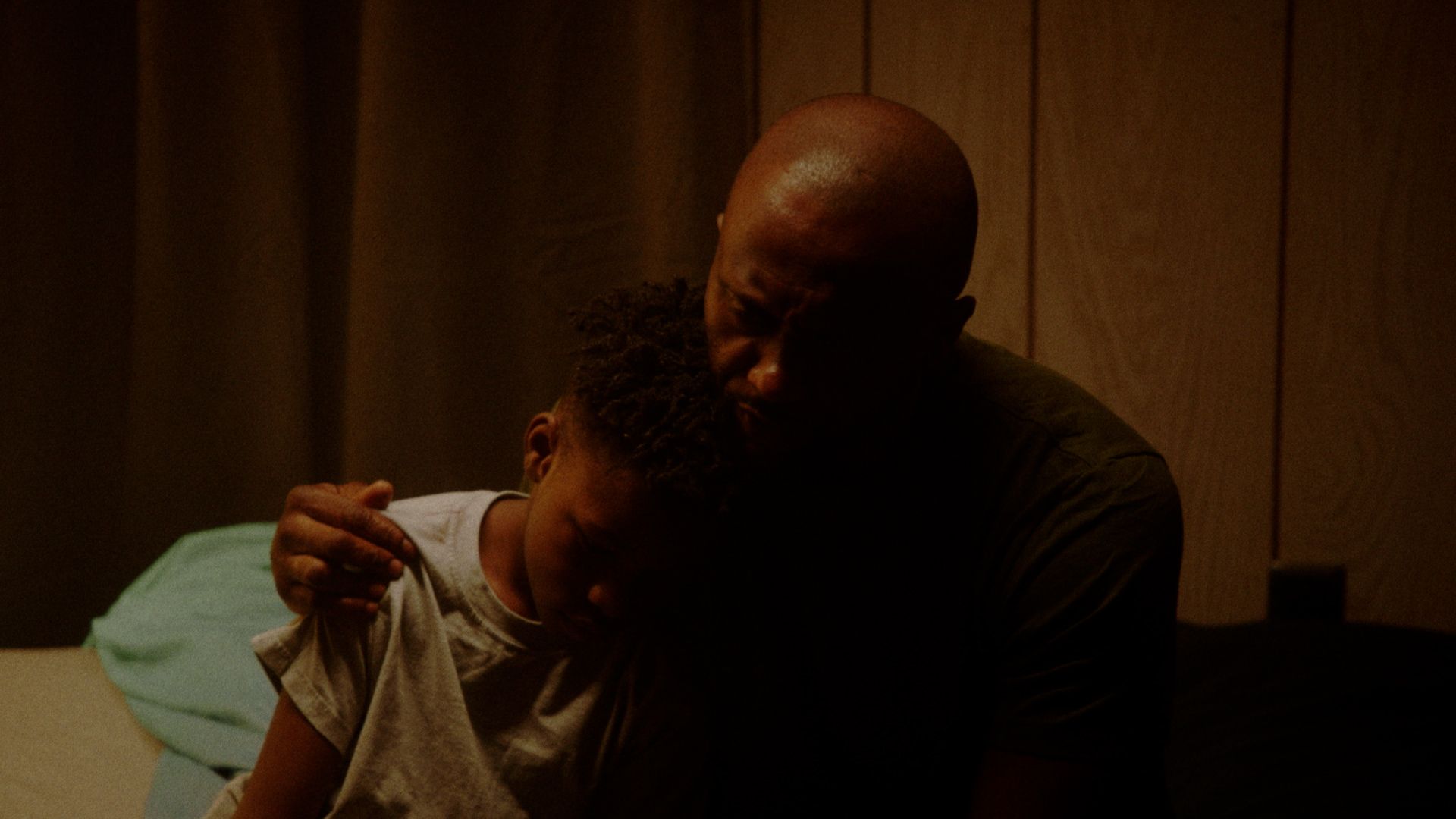 A father holds his son on the edge of a bed in a dim, wood-paneled room.